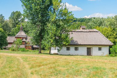Kırsal Orta Avrupa köyü, topraksı tonlar, kiremit çatılı ahşap binalar, ön planda ağaç, çimen, toprak yamalar, birden fazla pencereli daha büyük yapı, mavi gökyüzü, görünür insan yok, kıç tarafı geç.