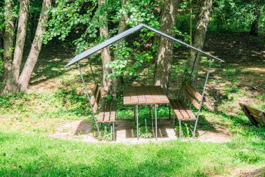 Orta Avrupa 'da yemyeşil ağaçlar, kahverengi ahşap ve çelik yapı ve yumuşak, hatta hafif çelikle çevrili antik bir köy ortamındaki eğimli metal bir barınağın altında Rustik ahşap piknik masası.