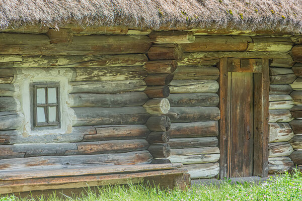 Выветриваемая бревенчатая хижина в древней среднеевропейской деревне, с центральным окном с несколькими стеклами, соломенной крышей и темной деревянной дверью