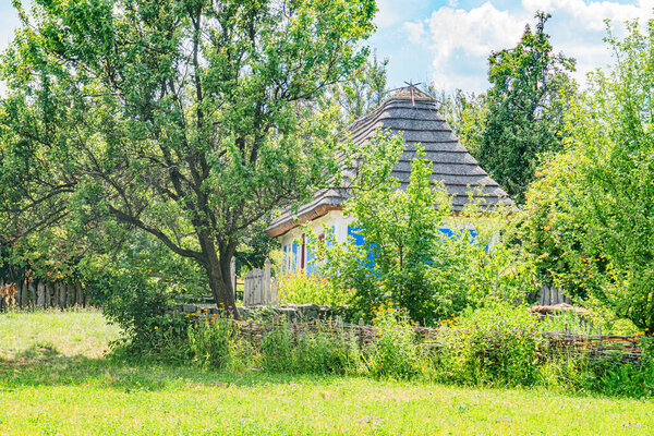 Древний соломенный коттедж среди зелени в среднеевропейской деревне, частично скрытая каменная стена или забор с левой стороны, покрытая травой земля Яркий дневной свет, яркие зеленые листья,