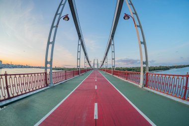 Bir kemer dizaynlı asma köprünün gün doğumu veya günbatımı görüntüsü, durgun sulara yayılmış, yemyeşil ağaçlarla çevrili köprü yolu boyunca kırmızı bir şeride sahiptir ve sıcak, nötrra ile betimlenmiştir.