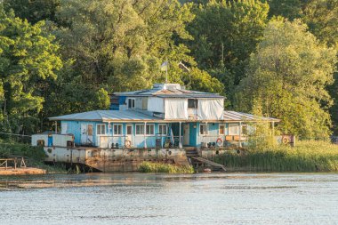 İki katlı, iskelede mavi bir ev, sakin su, balkon, perdeli pencereler, küçük tekne, arka planda ağaçlar, sabahın erken saatlerinde aydınlatma, doğal ortam, gerçekçi stil.