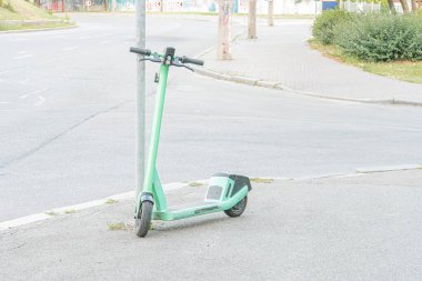 Şehir sokak sahnesinde kaldırıma park etmiş canlı yeşil elektrikli scooter, siyah tekerlekler ve gidonlar, doğal gündüz ışığı, sessiz renkler, benzersiz bir perspektif.