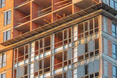 Alacakaranlıktaki bir inşaat sahasında uzun ve çok katlı bir bina sergileniyor. Yapı, sıralı kırmızı tuğla ve cam cephe içeriyor. Işıklar doğadan doğaya dönüşüyor.