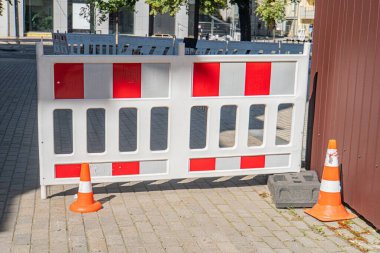 Tuğla döşeme üzerinde kırmızı, beyaz ve sarı dikey bariyerlerin ve turuncu konilerin yer aldığı kentsel alanda inşaat alanı güvenliği var.