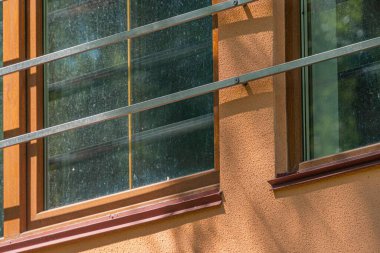 Modern bir şehir binasının köşesine yakın çekim, büyük kırmızı çerçeveli bir pencere, çoklu camların içindeki yeşillikleri ortaya çıkarıyor, güneş ışığının iç ve beyaz duvarlarda karmaşık desenler oluşturmasına olanak sağlıyor.