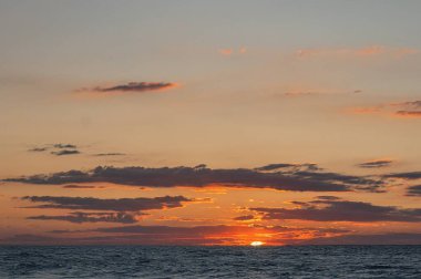 Büyük bir su kütlesi üzerinde gün batımı sahnesi, sıcak renkler turuncu, pembe, dağınık bulutlar, kısmen kararmış güneş, su yansıması, berrak ufuk, insan yapısı yok, gerçekçi bir tarz.