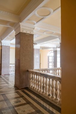 Eski bir Avrupa şehrinde sıcak tonlu, iyi ışıklandırılmış koridor, klasik mimari, uzun sütunlar, dekoratif pervazlar, kemerli giriş, ince desenli koyu fayanslı döşemeler, karmaşık parmaklıklar