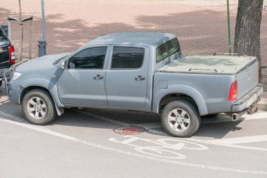 Koyu gri Toyota kamyonet şehir caddesinde park halinde, renkli arka pencere, yan profil görüntüsü, yedek lastiğe monte edilmiş, beyaz boyanmış çizgiler, ağaçlar, işaret direği, bulutlu gündüz aydınlatması, insan yok.