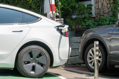 Bir elektrikli araç şarj istasyonunda gün ışığında iki elektrikli araba, biri Tesla Model S olarak tanımlanabilir ve diğeri belirsiz bir makemodel, sof ile doğal aydınlatma ile fişe takılır.