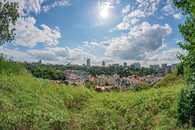 Yüksek görüş açısına sahip şehir manzarası, güneş ışığı altında verimli yeşillik ve dağınık bulutlar tarafından çerçevelenmiş konut ve ticari binaların karışımını gösteriyor.