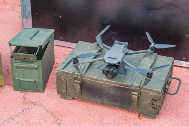 Askeri bir dron açık renkli bir yüzeye yakın iki yeşil depolama konteynırının üzerinde duruyor. İnsansız hava aracı parlak ve koyu renklidir, daha hafif pervaneleriyle zıt yöndedir. Konteynırlar sağlamdır, possi 'dir.