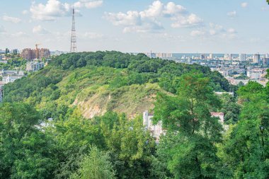 Yeşillik, dağınık ağaçlar ve uzak bir şehir manzaralı panoramik manzara. Önemli bir yapı, üssün etrafında görünen bir iletişim kulesi yolu gibi görünüyor.