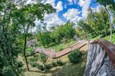 Yükselmiş, kıvrımlı bir metal korkuluk kentsel bir ortamdan yemyeşil bir parkın görüntüsünü sunar. Tırabzanlar yoğun yaprakların arasından parçalanmış bulutlu bir gökyüzünün altında kıvrılan bir yola çıkar.