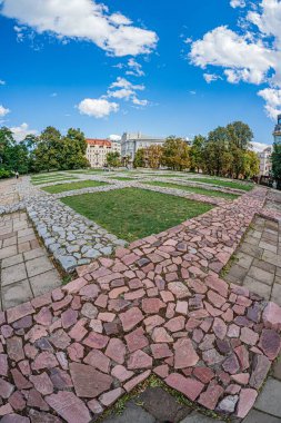 Eski bir Avrupa şehir bahçesinin yüksek açılı manzarası kırmızı tuğla patika, yemyeşil ağaçlar ve canlı yapraklar arasında beyaz bir mermer çeşmeye götürür.