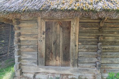 Yıpranmış ahşap bir kulübe, dikey kütükler ve sazdan çatı ile inşa edilmiş küçük bir veranda alanı, ahşap bir lintel ile güçlendirilmiş bir giriş ve muhtemelen bir havalandırma kazancı vardır.