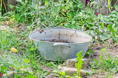 Antik Orta Avrupa köyü Toprak zeminde büyük metal kazan, bitkiler ve yabani otlarla çevrili Kısmen saklanmış ahşap çit yapısı Gündüz vakti doğal ışık bırakmıyor