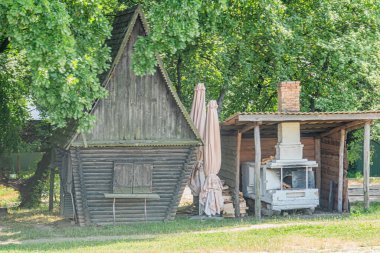 İki ahşap kulübesi olan Rustik Orta Avrupa köyü sahnesi, biri yeşillik ile gizlenmiş, diğeri önde gelen beyaz araç ve metal silindir ile önde gelen ahşap bank nea