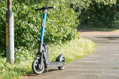Bulutlu bir gün sahnesi, ıslak beton caddede, yeşil çimenler ve ağaçlarla çevrili mavi ve beyaz bir scooter 'la yüksek bir açıdan çekiliyor ve net bir görüş sağlıyor.