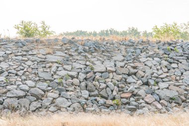 Açısal, düzensiz şekilli gri taşlar engebeli bir desen oluşturarak duvarımsı bir yapı oluşturuyor. Şehir arazisinde, hareketsiz bitki örtüsü ve uzak doğa çalıları, ağaçlar S