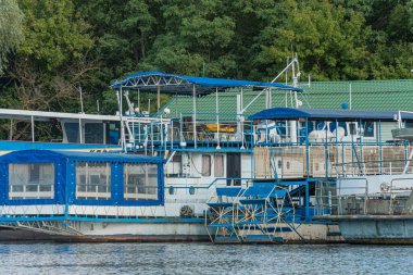 Rıhtımda boş bir tekne, doğal bir ortamda pencereleri, tırabzanları ve can simidi olan sessiz mavi bir yapı.