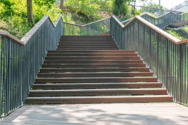 Çağdaş metal merdiven yemyeşil bir şehir ortamında dayanıklı metal paneller güneş ışığını yansıtıyor, ahşap bir zeminden yükselen yüksek bir platforma yükseliyor.