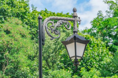Açık şehir ortamında metalden dekore edilmiş eski bir sokak lambasıyla yeşil ve doğal gün ışığının yer aldığı eski bir Avrupa şehri manzarası.
