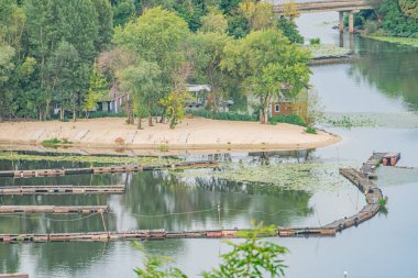 Küçük tekne çok katlı bir rıhtıma ya da nehir kenarındaki eve kenetlendi, bitki örtüsü ve su bitkileriyle sakin bir nehir, toprak rengi paleti, yumuşak bulutlu gökyüzü, gerçekçi bir tarz.