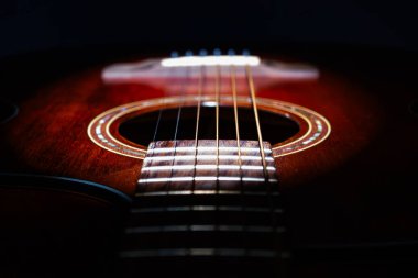 Eski bir ahşap akustik gitarın karanlık arka plana karşı yakın çekimi, teller dikey çalışıyor, akort mandallı başlık, sıcak aydınlatma, dokulu detaylara odaklanan hafif açı görünümü