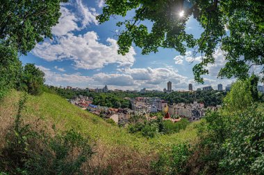 Canlı bir kentsel doğa bileşimi olan izleyici yeşil bir gölgelikten şehri gözlemler, benekli güneş ışığı gölgeleri ile şehir mavi gökyüzünün altında beyaz ve gri renkli binaların bir karışımıdır.