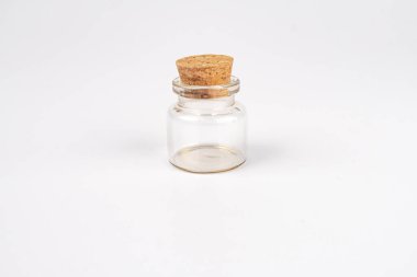 Açık renkli cam kavanoz, muhtemelen bal, koyu kahverengi tıpa tıpası ve metal halka ile mühürlenmiş, açık gri zemin üzerine yayılmış ışık ince gölgeler oluşturur.