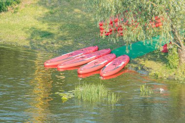 Kırmızı kanolar sakin su boyunca bağlanmış, muhtemelen bir nehir ya da göl. Kıyı şeridinden görünen yeşil yapraklar. Bulutlu gökyüzü yumuşak ışık sağlıyor.