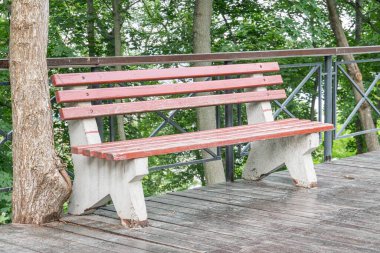 Şehir parkında ya da bahçe düzeninde yükseltilmiş yeşil bir platformda sıcak kahverengi ahşap bir bank var. Platformun metal bir korkuluğu var ve bulutlu gökyüzünün altında yemyeşil yemyeşil ile çevrili.