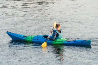 Çok renkli kano, sarı şerit ve kürek, mavi can yeleği, yeşilimsi mavi su, hareketli kürek, yer işareti yok, bulutlu gün ışığı, belgesel tarzı, p hattının üstünde.