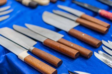 Koyu renk kumaş üzerinde ahşap sapları olan çeşitli bıçaklar, değişik uzunluklar ve tasarımlar, sıcak tonlarla zıt metalik bıçaklar.