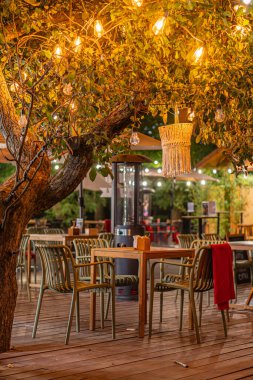 Kırmızı masa örtüleri, tel lambaları ve şehir merkezinde büyük bir ağacı olan gece açık hava yemek alanı.