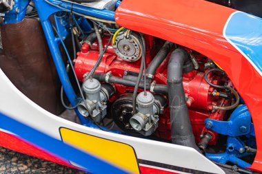 Bir atölyede veya yarışta kırmızı ve mavi bileşenleri gösteren klasik bir motorun yakın çekim görüntüsü. Dikkat çekici detaylar, hortumlar, tüpler, ısıya dayanıklı kırmızı parçalar.