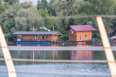 Ukrayna 'nın başkenti Kyiv' de sakin bir göl eğimli çatıları ve balkonları olan iki ahşap ev, Ukrayna, Kyiv, 06-22-2025 ön tarafındaki büyük yeşil ağaçların hüküm sürdüğü yakınlardaki mavi bir tekne.