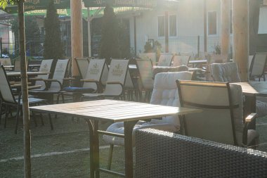 Yemek sandalyeleri, masalar, ahşap döşemeler, tarafsız mobilyalar, çitler, bina, yeşillik, yumuşak ışıklandırma, öğleden sonra geç saatlerde kırsal kontrastlar, sakin bir Kyiv, Ukrayna 06-26-2025