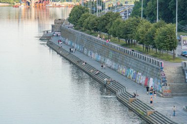 Alacakaranlıkta Kyiv rıhtımı, beton deniz kenarında canlı grafiti, sakin yansıtıcı sular, demirli kayıklar, yoğun ağaçlar, berrak gökyüzü, uzaktaki Kyiv, Ukrayna 07-05-2025