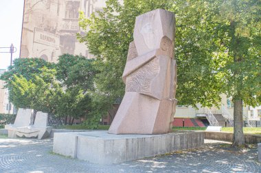 Şehir parkındaki beton kaide üzerine açısal taş heykel soyut, geometrik şekiller, kırmızımsı kahverengi renkler, yemyeşil ağaçlar, yumuşak dağınık Kyiv, Ukrayna 07-06-2025