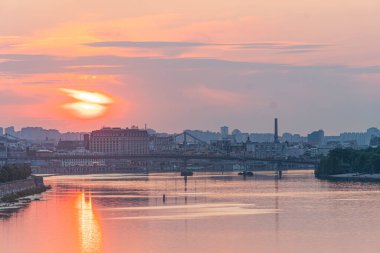 Gün batımında sakin bir nehir, turuncu gökyüzü yansıması, küçük dalgalar, rüzgarlı, nehir kıyısı ağaçları, şehir manzarası, eski ve yeni yapıların karışımı, sokak lambaları veya sokak direklerinin karışımı, Kyiv, Ukrayna 07-06-2025