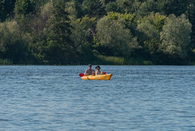 Sakin göl manzarası, gündüz aydınlığı, iki kişi, sarı kano, kırmızı yelkenli, yemyeşil, açık hava eğlencesi, doğa keyfi Kyiv, Ukrayna 07-12-2025