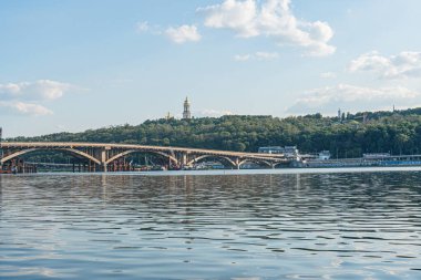 Köprüsü olan sakin bir nehir, uzak inşa kulesi, dağınık bulutlu açık mavi gökyüzü, birden fazla kemer köprüsü, karşı kıyılara gerçekçi bir şekilde bağlanan doğal I Kyiv, Ukrayna 07-12-2025