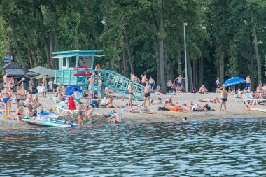 Kyiv, Ukrayna 'da plaj sahnesi güneşlenen insanlar, şemsiyeler, cankurtaran kulesi, kumlu sahil, mavi su, botlar, canlı renkler, ağaçlar, yeşil, gökyüzü, balık gözü efekti, Kyiv, Ukrayna 07-12-2025