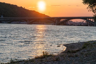 Ukrayna 'nın başkenti Kyiv' de gündoğumunda geçen özet nehir sahnesi. Sıcak renkler, kısmen görünür güneş, uzun gölgeler, sakin nehir, kemerli köprü, durgun su yüzeyi, çimen, çalılık.,