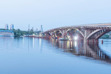 Alacakaranlıkta suyun üzerinde birden fazla kemer bulunan köprü, dahili ışıklarla aydınlatılıyor. Ön planda vinçlerle görülebilen gemiler inşa ediliyor. Kyiv, Ukrayna 06-01-2025 'i yansıtan sakin su.