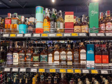 Bir süpermarkette, çeşitli viski, votka, cin, dikey ve yatay olarak düzenlenmiş etiketli şişelerle dolu metalik alkol rafı görüntüsü.