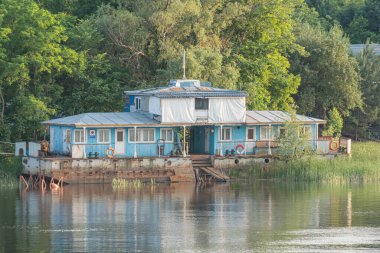 Suların kenarındaki mavi iki katlı ev, güverte, beyaz pencere çerçeveleri, ahşap iskele, yemyeşil, sakin su, sakin kırsal alan, öğleden sonra Kyiv, Ukrayna 06-05-2025