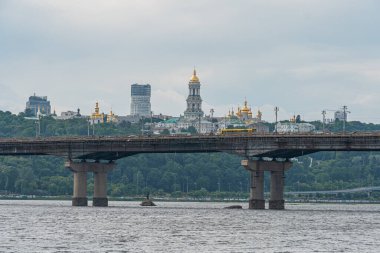 Ukrayna 'nın başkenti Kyiv' in havadan görünüşü, Dinyeper Nehri üzerinde farklı rıhtımları olan bir asma köprü barındırıyor. Uzakta ise altın rengi Kyiv, Ukrayna 06-14-2025 'i de içeren binalar bulunuyor.